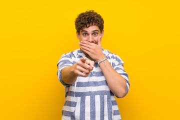 Blonde man over yellow wall pointing with finger at someone and laughing