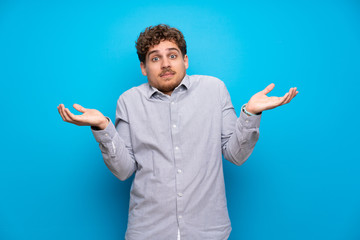 Blonde man over blue wall having doubts while raising hands