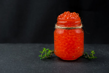 Glass jar with red caviar and fresh thyme on dark  background. Healthy food.