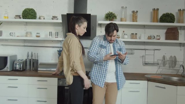 Young wife suspecting using smartphone husband in betrayal while seeing him chatting online in domestic kitchen. Jealous woman taking away cellphone to make sure beloved man texting to mistress.