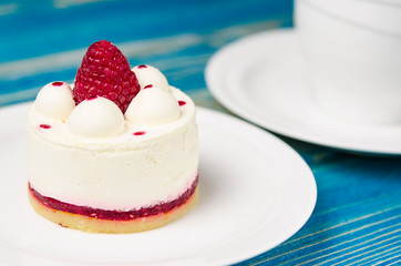 White airy cake with raspberry layer and raspberry berry lies on a white round plate next to a white cup, which stands on a blue table