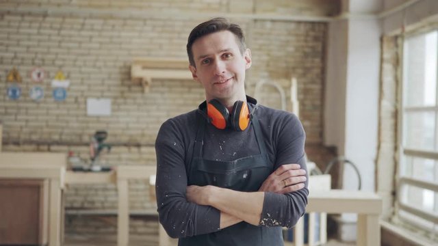 Tilt up shot of handsome carpenter posing at workbench crossing arms and looking at camera, protective earmuffs around his neck