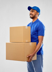 mail service and shipment concept - happy indian delivery man with parcel boxes in blue uniform over grey background