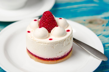 White airy cake with raspberry layer and raspberry berry lies on a white round plate next to a white cup, which stands on a blue table