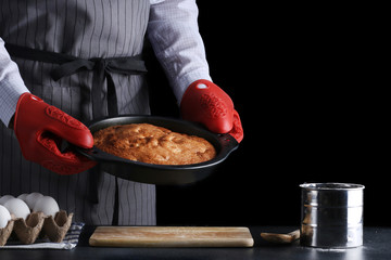 man in apron holding freshly baked pie on dark background. recipe od pie or cake