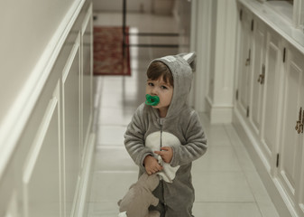 A cute toddler with a very funny pajamas and a pacifier, goes down the hall with a bunny