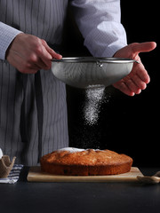 chef pouring powdered sugar on tasty pie on dark background. ched processing pie or cake