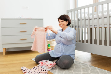 motherhood, pregnancy and nursery concept - happy pregnant middle-aged woman reviewing baby clothes...