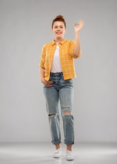 people concept - smiling red haired teenage girl in checkered shirt and torn jeans showing ok hand sign over grey background