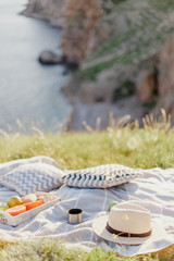 Picnic on the grass on mountain over sea view on sunny summer day. Relaxing on nature. Tray with fruits, cup of tea, straw hat and pillows lying on plaid or blanket.