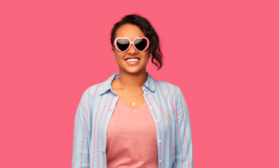 summer, valentine's day and eyewear concept - happy african american young woman in heart-shaped sunglasses over pink background