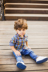 Naklejka premium Portrait of handsome little toddler boy playing at summer city park. Calm child with beautiful curly brown hair and big eyes posing for photo sitting on wooden steps. Vertical color photography.