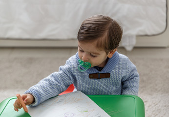 A beautiful child with pacifier paints with crayons on a small desk
