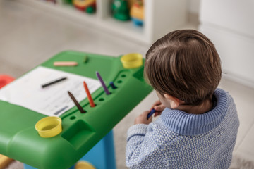 A little blond boy with a pacifier and dressed in blue, paints with crayons on a sheet, while he is...