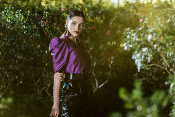 Pensive girl in purple blouse and leather pants posing in botanical garden