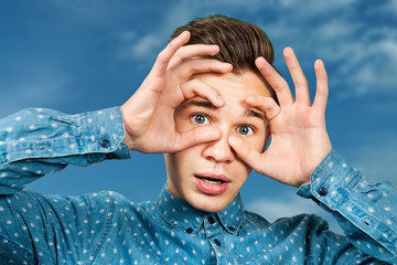 Portrait, young, guy, dressed, blue, shirt., man, open, his, eyes, wide, with hands, fingers, and mouth. Man on blue sky background
