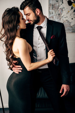 attractive woman in black dress holding tie of passionate man standing in suit
