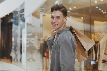 Handsome happy young man smiling to the camera over his choulder, carrying shopping bags at the mall. Attractive male customer enjoying sale at the shopping mall. Consumerism concept