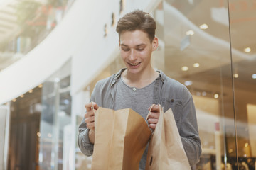 Charming young man smiling, looking inside his shopping bag, enjoying sale at the mall. Cheerful male customer shopping at the mall. Retail, spending money, leisure concept