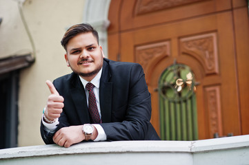 Stylish indian businessman in formal wear leaning on a railing against door in business center. He happy man and show thumb up.