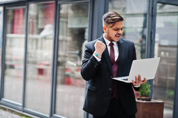 Fototapeta premium Stylish excited happy indian businessman in formal wear with laptop on hands standing against windows in business center. Gesture of the winner!