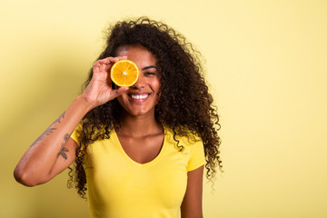Portrait of young woman holding an orange against her eye. Female model looking at camera over yellow background.
