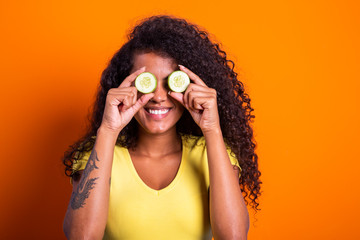 Fun portrait of beautiful black african model holding a cucumber slice to her eye, rejuvenating skin care regime treatment facial beauty concept.
