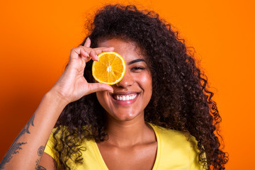 Portrait of young woman holding an orange against her eye. Female model looking at camera over yellow background.