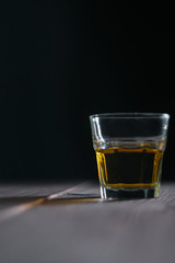 Vertical photo of a whiskey glass on the right on a wooden table with a dark background behind. Table in front of a glass in defocusing
