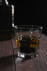 Vertical contrast photo of a glass of whiskey and stones standing in the foreground on a wooden table. In the background is a fragment of a bottle.
