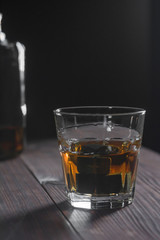 A glass of whiskey and stones stands in the foreground on a wooden table in the background a fragment of a bottle