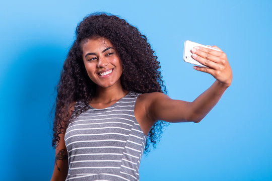 Closeup portrait of smiling young attractive African brazilian woman holding smartphone, taking selfie photo on the blue background - Imagem