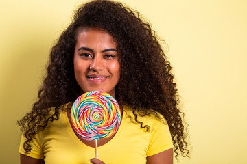 Smiling curly african girl in bright yellow t-shirt holding candy lollipop. Young cheerful woman...