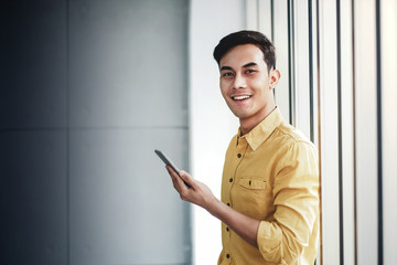 Portrait of Happy Businessman Standing by the Window in Office. Using Smartphone and Smiling. Looking at Camera