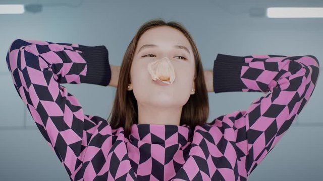 Portrait of a young playful brunette Asian girl standing and hands behind her head blowing bubblegum candy bubbles and chewing gum sticking to her nose. The woman looks at the camera. 4k. Slow motion