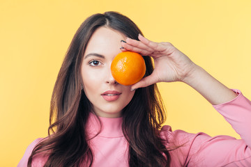 Beautiful young woman holding orange in front of eye isolated on yellow background