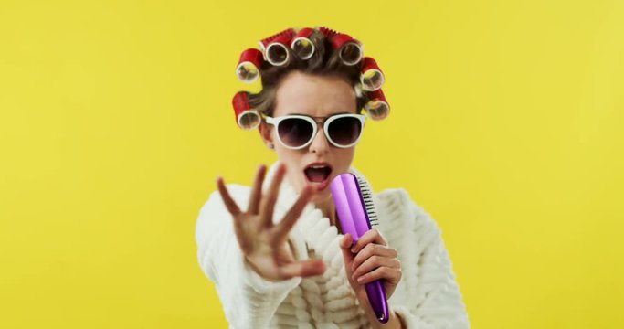 Portrait shot of the funny Caucasian woman ib the bathing robe, curlers and sunglasses singing in the hair comb on the yellow background.