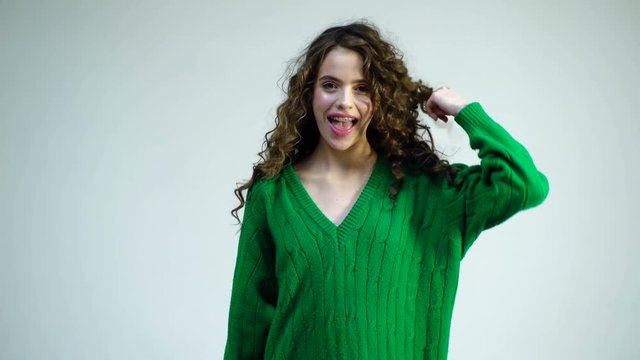 Fashion woman in green sweater. Cheerful beautiful girl with curly hair, braces on her teeth, wraps hair on a finger, smiling broadly with her mouth open. Concept fun, happiness, laughter.