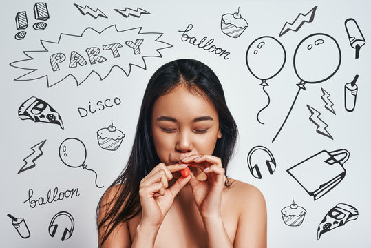 Party time! Beautiful asian girl with long hair is trying to blow a balloon while standing against grey background with party theme doodles on it.