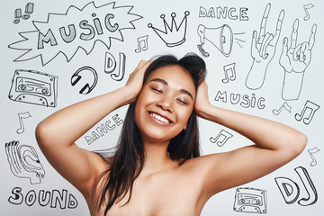 Enjoying my favourite song. Smiling cute asian woman touching her hair and relaxing with closed eyes while standing against grey background with music theme doodles on it.