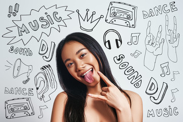 Music is my freedom. Joyful young age asian woman gesturing and sticking out tongue while standing against grey background with music theme doodles on it.