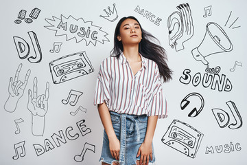 Fototapeta premium Relaxing with my favourite music...Young charming asian woman in stylish wear is looking at camera while standing against grey background with music theme doodles