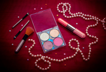 Makeup cosmetic kit – eye shadows, highlighter, lip gloss and blush, brushes and pink string of pearls on a dark red background