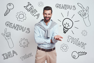 Great idea! Charming bearded man in a blue shirt is smiling at camera while standing against grey background with different doodle illustrations on it.
