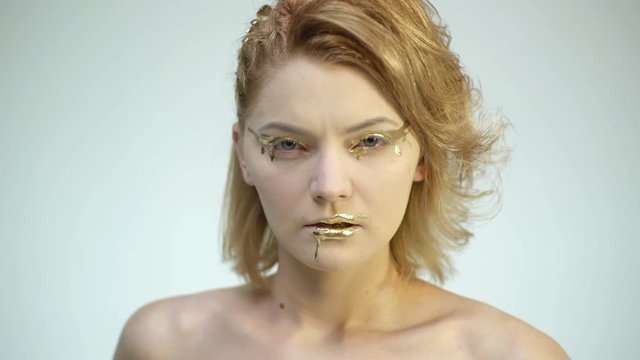 High fashion body art. Woman with golden skin and lips. Glamour shiny professional makeup. Gold Paint smudges drips from woman face and lips. Golden liquid drops on girl's mouth.