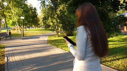 girl with tablet in light business suit goes to work. beautiful businesswoman walks along alley in park with tablet in her hands.