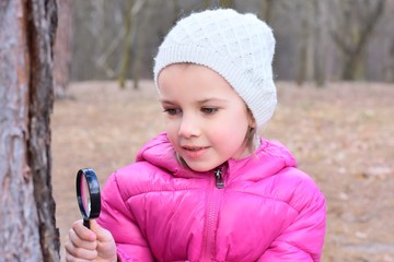 Pretty caucasian girl with funny face in pink coat looking through a magnifying glass pine bark with rough texture. Portrait of a beautiful white girl studying the environment with a magnifier lens