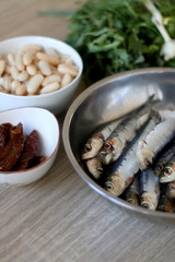 Fresh sardines, white beans, dried tomatoes and chicory on a wooden table. Ingredients for a meal. Selective focus.