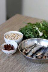 Fresh sardines, white beans, dried tomatoes and chicory on a wooden table. Ingredients for a meal. Selective focus.