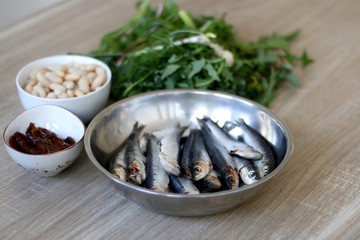 Fresh sardines, white beans, dried tomatoes and chicory on a wooden table. Ingredients for a meal. Selective focus.